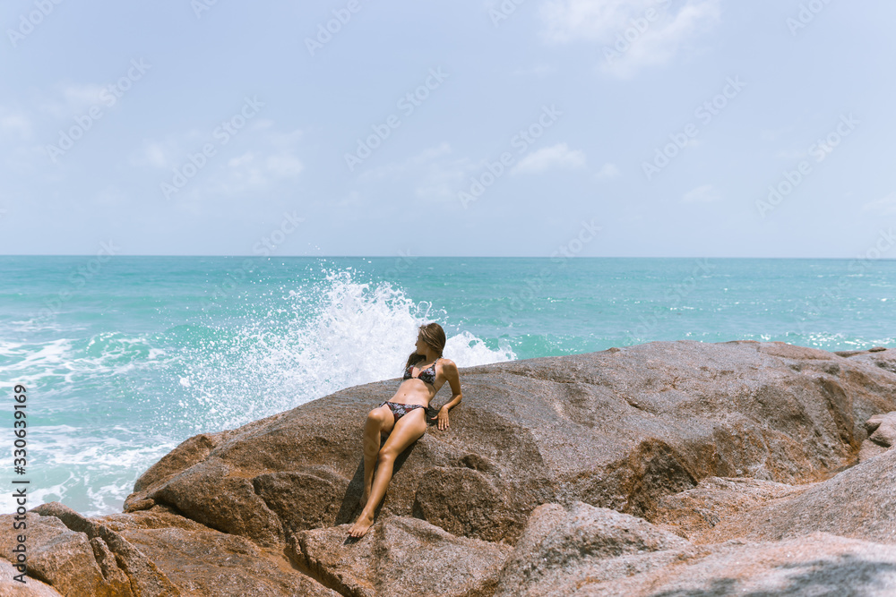 A beautiful girl in a swimsuit is standing on the beach on the rocks with the sea.Against her background,waves and splashes splash.The concept of a traveler and a good life in Thailand.Model