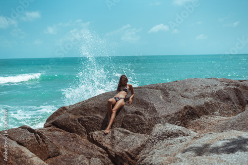 A beautiful girl in a swimsuit is standing on the beach on the rocks with the sea.Against her background,waves and splashes splash.The concept of a traveler and a good life in Thailand.Model