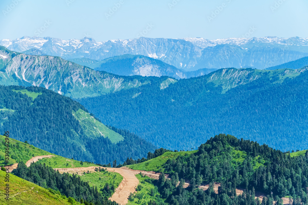 Green mountain ridges, surrounded by high mountains. Snow-capped mountain peaks on the horizon. Dirt road on mountain range. Layers of mountains in the haze. Krasnaya Polyana, Sochi, Caucasus, Russia
