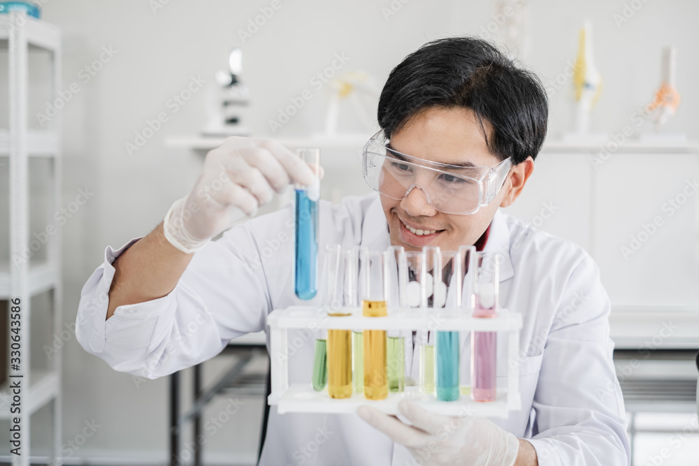 A male scientist with black hair wearing white coat and protective glassware sitting smiling looking at test tube and solutions in white laboratory room. A male scientist with black hair wearing white coat and protective glassware sitting smiling looking at test tube and solutions in white laboratory room.