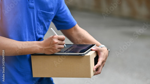 Cropped image of delivery man using a stylus pen and computer tablet for checking address of parcel location while standing outdoors somewhere close to customer house.