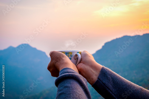 Hand of traveler holding coffee cup at sunrise and mountain range at front, freedom traveling lifestyle concept