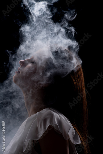 The silhouette of a young beautiful girl in a white dress in a cloud of smoke, a cloud of thick smoke descends on the woman 's face