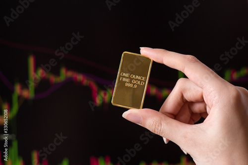 The hand holds a gold bar on the background of a laptop on which there is a trading price chart. One ounce of gold in hand. Trading gold on the stock exchange and Forex.