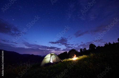 Horizontal snapshot of an illuminated tent and campfire under incredible starry sky  summer time evening in the mountains  astrophotography  copy space. Night camping