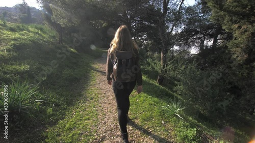 Young woman walks on path among trees and grass on sunny day, looks back at camera and smiles, gimbal following photo