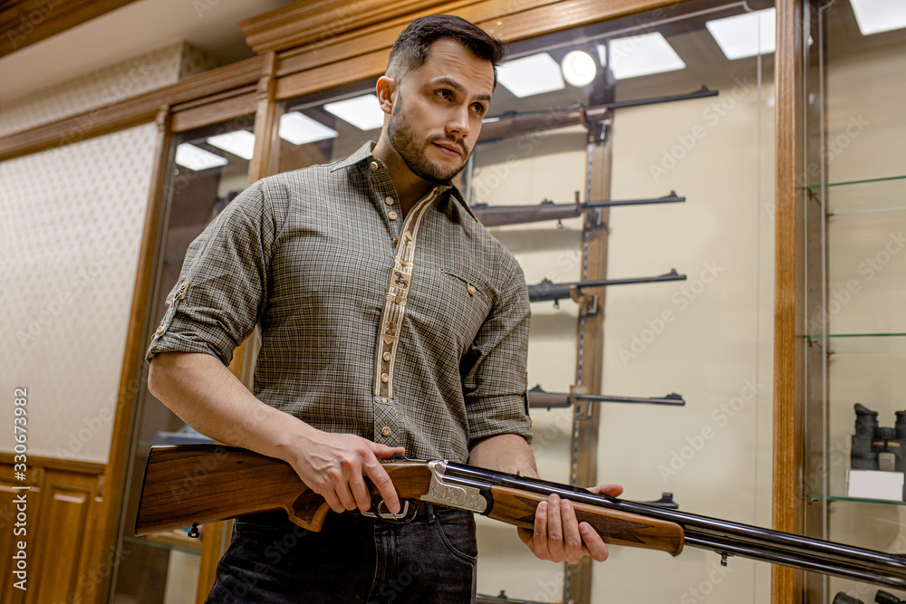 handsome caucasian guy looking for good weapon rifle in guns store. man choose and studying different rifles. he hold one weapon in hand and want to buy it for hunting