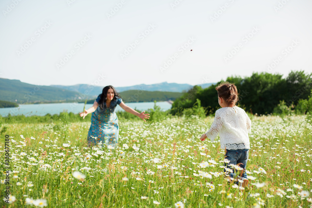Fototapeta premium Active kid dressed jeans and t-shirt running and have fun to pregnant mother on the nature green field with chamomiles. Healthy lifestyle concept