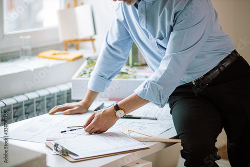 cropped adult man in blue shirt and trousers sit at table in office, unrecognizable architect working with drawings next to window