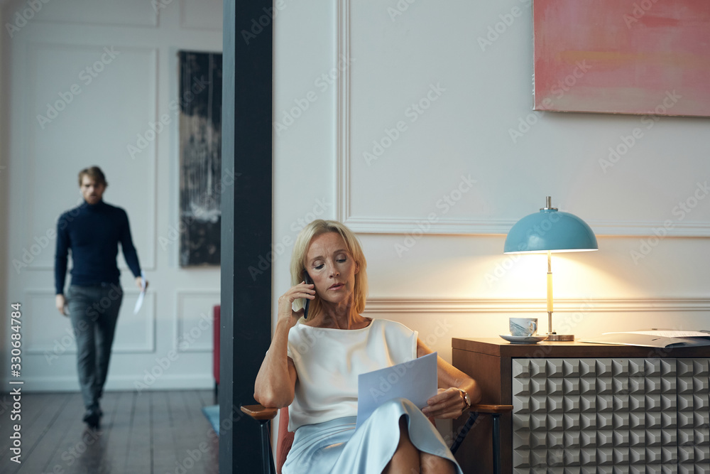 Portrait of successful mature woman speaking by phone and holding documents while sitting in elegant interior with man in background, copy space Portrait of successful mature woman speaking by phone and holding documents while sitting in elegant interior with man in background, copy space