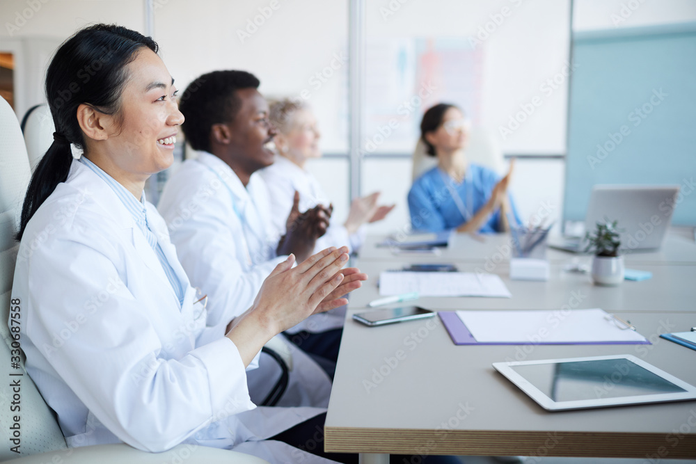 Side view at multi-ethnic group of doctors applauding while sitting at table during medical conference, focus on smiling Asian woman in foreground, copy space