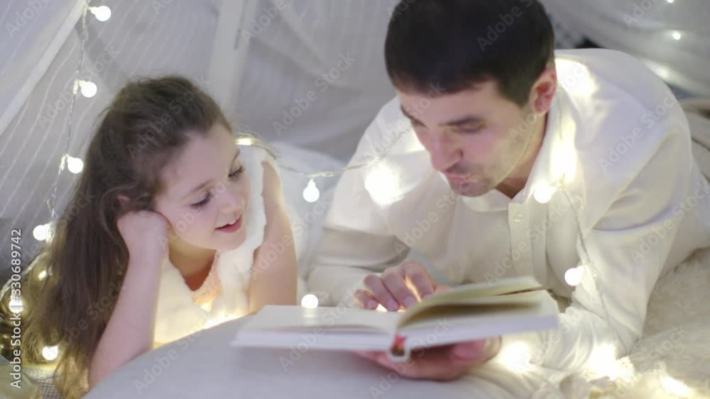 Father in White Shirt and Little Girl in a White Dress Lies on the Carpet and Reading Book Together in Christmas Decoration. New Year Family Holiday. The Concept of a Parenthood, Family. Father in White Shirt and Little Girl in a White Dress Lies on the Carpet and Reading Book Together in Christmas Decoration. New Year Family Holiday. The Concept of a Parenthood, Family.