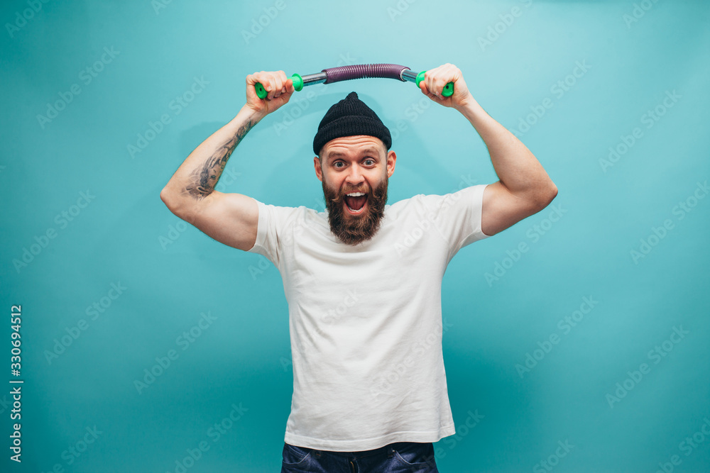 Naklejka premium Handsome bearded hipster guy exercising with expander. Crazy sports man emotions. Guy with beard wearing casual beanie and white t shirt.