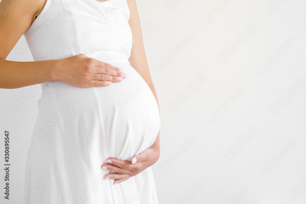 Young pregnant woman in white dress touching big belly with hands. Baby expectation. Pregnancy time - 37 weeks. Closeup. Empty place for text on gray background. Side view. Young pregnant woman in white dress touching big belly with hands. Baby expectation. Pregnancy time - 37 weeks. Closeup. Empty place for text on gray background. Side view.