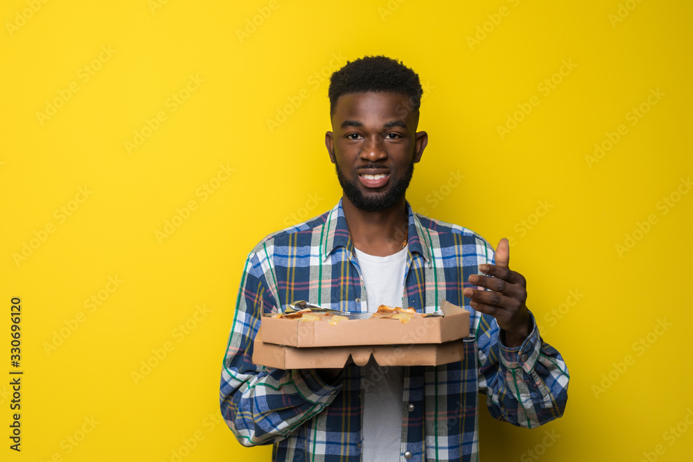 Afro american man eating slice of pizza over isolated yellow background Afro american man eating slice of pizza over isolated yellow background