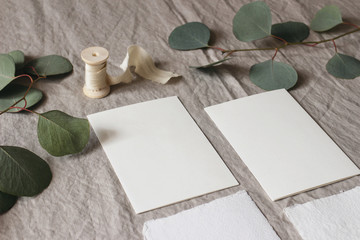 Wedding stationery mock-up scene. Blank greeting cards on linen tablecloth background with green eucalyptus tree leaves and branches and silk ribbon. Feminine still life composition. High angle view.