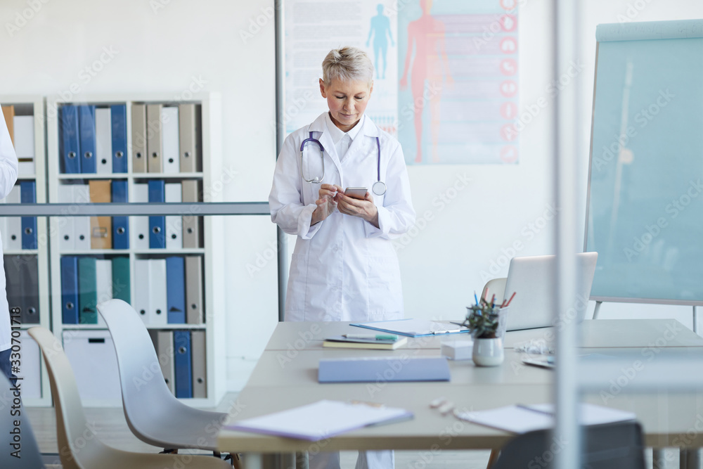 Portrait of mature female doctor using smartphone while standing at table in conference room in medical office interior, copy space