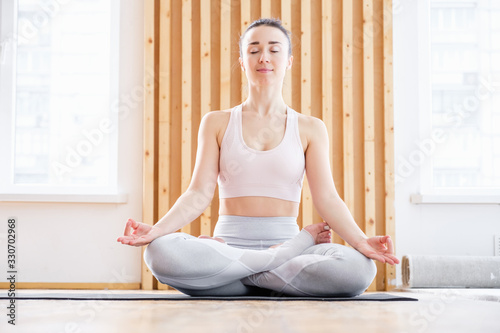 Healthy pacified young woman in tracksuit practices yoga in lotus position and mudra gesture on rug on floor on white brick wall. Healthy respiratory and nervous system concept. Advertising space