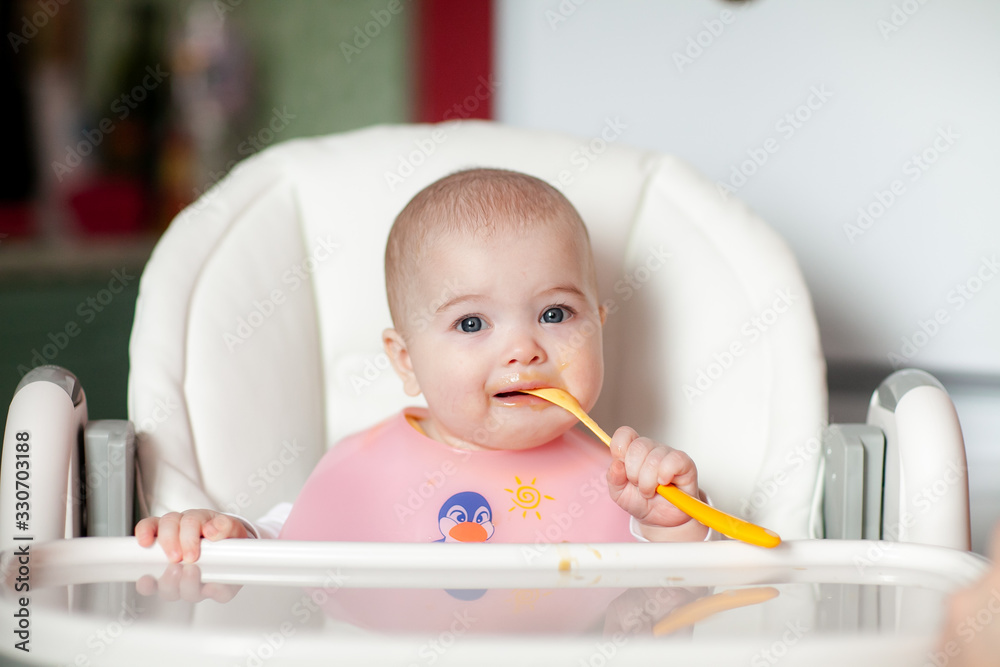 Cheerful baby child eats food itself with spoon. Portrait of happy kid girl in high chair. Cheerful baby child eats food itself with spoon. Portrait of happy kid girl in high chair.