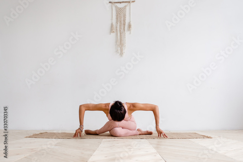 Young attractive woman practicing yoga in bright yoga class on wooden vintage floor, sitting in variation of Gomukasana exercise with forward bend, Cow Face pose, working out, wearing pink sportswear