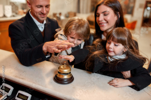 Fully enjoy your stay. Happy family checking in hotel at reception desk. A son and a daughter are ringing a service bell together with parents while standing at the hotel reception.