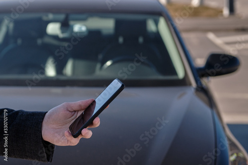A man in a coat holds a smartphone on the background of the car and controls it
