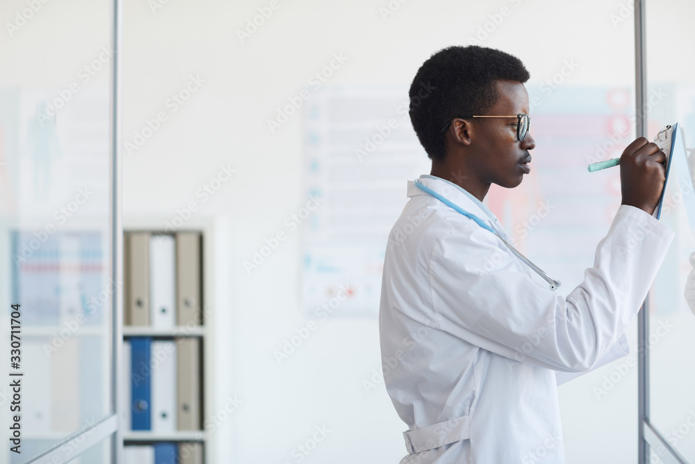 Side view portrait of young African-American doctor writing on clipboard while standing by glass wall in med clinic, copy space