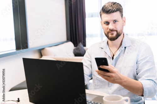Man with a beard and blue eyes working remotely with a laptop and a mobile phone in his hand