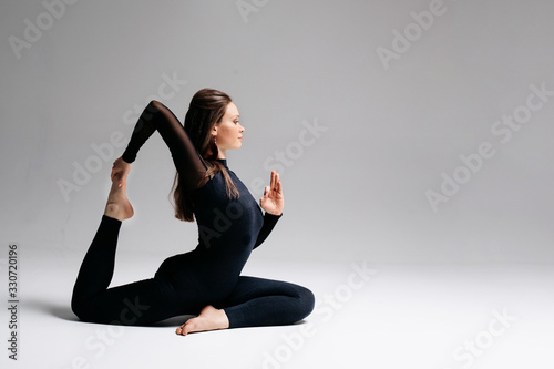 A beautiful girl of European appearance brunette sits in a difficult pose from yoga. The legs are twine, one leg is raised up, the hand holds the leg. Photo on a white background.