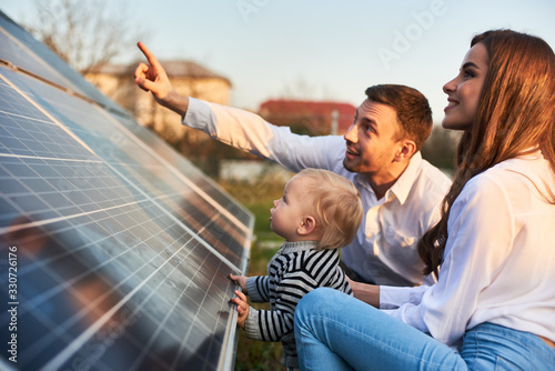 Man shows his family the solar panels on the plot near the house during a warm day. Young woman with a kid and a man in the sun rays look at the solar panels. photo