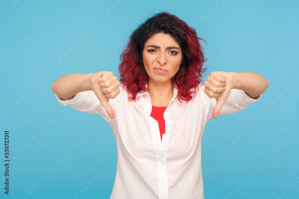 Fototapeta premium Dislike, bad review! Portrait of frustrated dissatisfied hipster woman with fancy red hair showing thumbs down, expressing disapproval, critique. indoor studio shot isolated on blue background