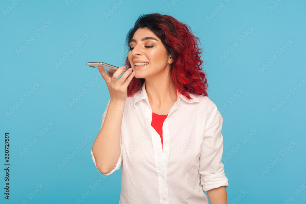 Innovative technologies. Hipster woman with fancy red hair asking voice assistant on cell phone, giving task, talking to artificial intelligence in mobile device. indoor studio shot, blue background Innovative technologies. Hipster woman with fancy red hair asking voice assistant on cell phone, giving task, talking to artificial intelligence in mobile device. indoor studio shot, blue background