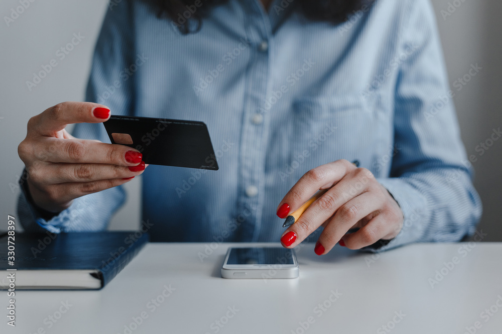 Closeup of woman's hands with red nails holding credit card and clicking mobile phone making payment online wearing blue shirt. Business, freelance, self-employment. Distance job, online work concept.