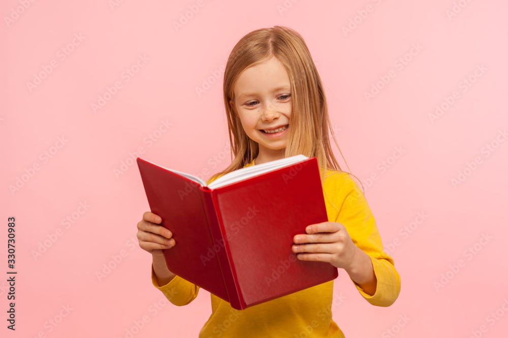 Smart positive cute little girl reading big red book and smiling happily, learning homework, being curious about school knowledge, education concept. indoor studio shot isolated on pink background Smart positive cute little girl reading big red book and smiling happily, learning homework, being curious about school knowledge, education concept. indoor studio shot isolated on pink background