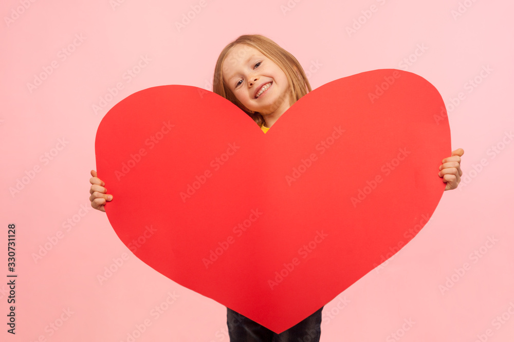 Portrait of sweet cheerful little girl peeking out of large red heart symbol and smiling to camera, congratulating on Mother's day, saying I love you. indoor studio shot isolated on pink background Portrait of sweet cheerful little girl peeking out of large red heart symbol and smiling to camera, congratulating on Mother's day, saying I love you. indoor studio shot isolated on pink background