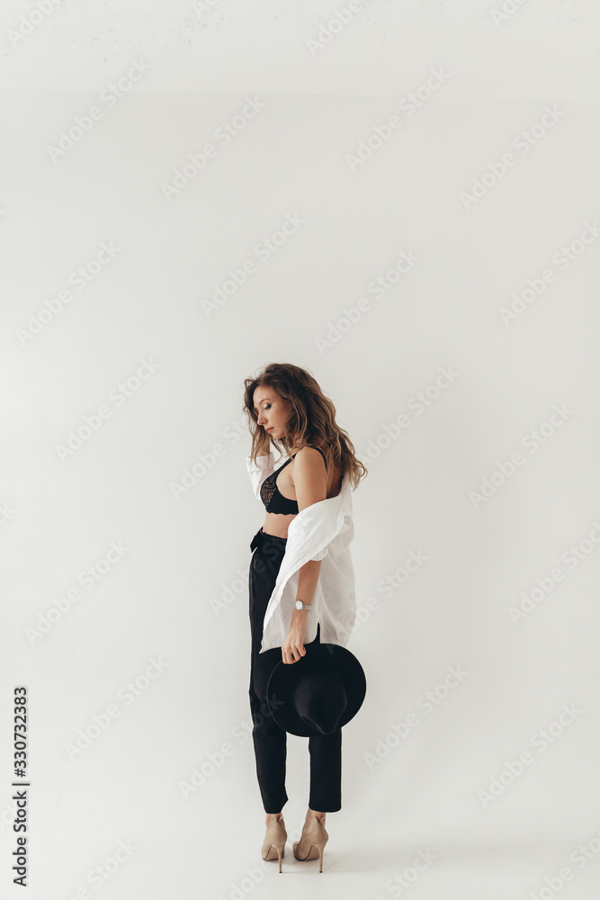 The concept of a trendy modern fashion trend shooting in a photo Studio , a beautiful sexy attractive gentle daring girl in a black hat, in black underwear, in a large shirt posing on a white backgrou The concept of a trendy modern fashion trend shooting in a photo Studio , a beautiful sexy attractive gentle daring girl in a black hat, in black underwear, in a large shirt posing on a white backgrou