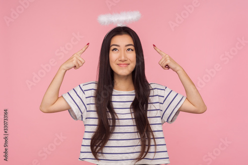 Portrait of shy modest asian girl with long brunette hair in striped t-shirt pointing at angelic halo over her head, showing nimbus and timidly smiling. indoor studio shot isolated on pink background