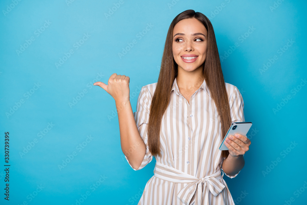 Sale. Photo of pretty attractive lady blogger hold telephone arms chatting friends directing thumb finger side empty space wear casual striped dress isolated blue color background Sale. Photo of pretty attractive lady blogger hold telephone arms chatting friends directing thumb finger side empty space wear casual striped dress isolated blue color background