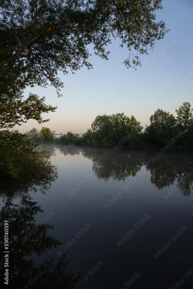 Beautiful morning river, a lake in light fog and the rays of the sun. Trees and bushes glow in the evening. Stock nature background for design.