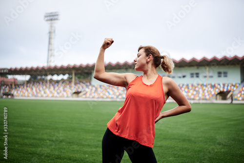 Three-quarter portrait of blond sportswoman, wearing orange top and black leggings, standing on stadium with her arms up, showing her muscles. Powerful fit strong woman, ready for fitness training