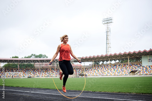 Full-length picture of sportswoman, jumping on jump rope on stadium. Young blond woman, wearing orange top and black leggings, doing fitness training in the morning in summer.