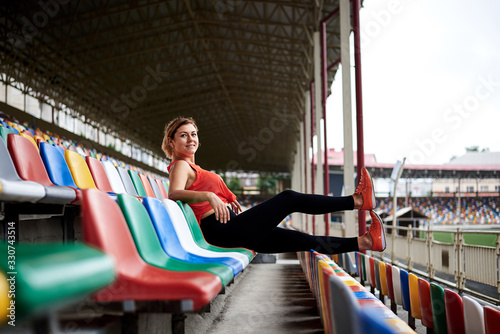 Young blond woman, wearing orange top, black leggings, orange sneakers, sitting on colorful seat chair on stadium, relaxing after training with her legs up on railing. Sportswoman on empty tribune.