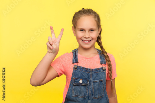 Portrait of positive adorable little girl with braid in denim overalls showing v sign with double fingers, gesturing victory, celebrating success. indoor studio shot isolated on yellow background