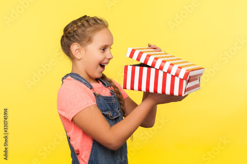 Portrait of glad curious little girl with braid in denim overalls peeking into gift box and laughing from happiness, enjoying best surprise, holiday bonus. studio shot isolated on yellow background photo