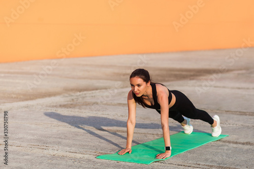Beautiful fit brunette woman in tight sportswear, black pants and top, practicing yoga, standing in Phalakasana higher plank pose, training muscles and strength. Health care, sport activity outdoor