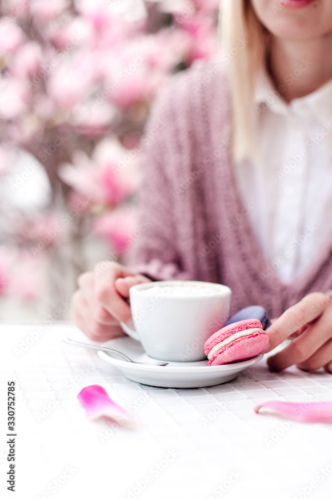 Young woman drinking coffee. Spring french cafe. Cup and pink macaroons in female hands. Girl enjoying morning breakfast outdoors in garden. Blooming magnolia flowers at city streets. Lifestyle moment Young woman drinking coffee. Spring french cafe. Cup and pink macaroons in female hands. Girl enjoying morning breakfast outdoors in garden. Blooming magnolia flowers at city streets. Lifestyle moment