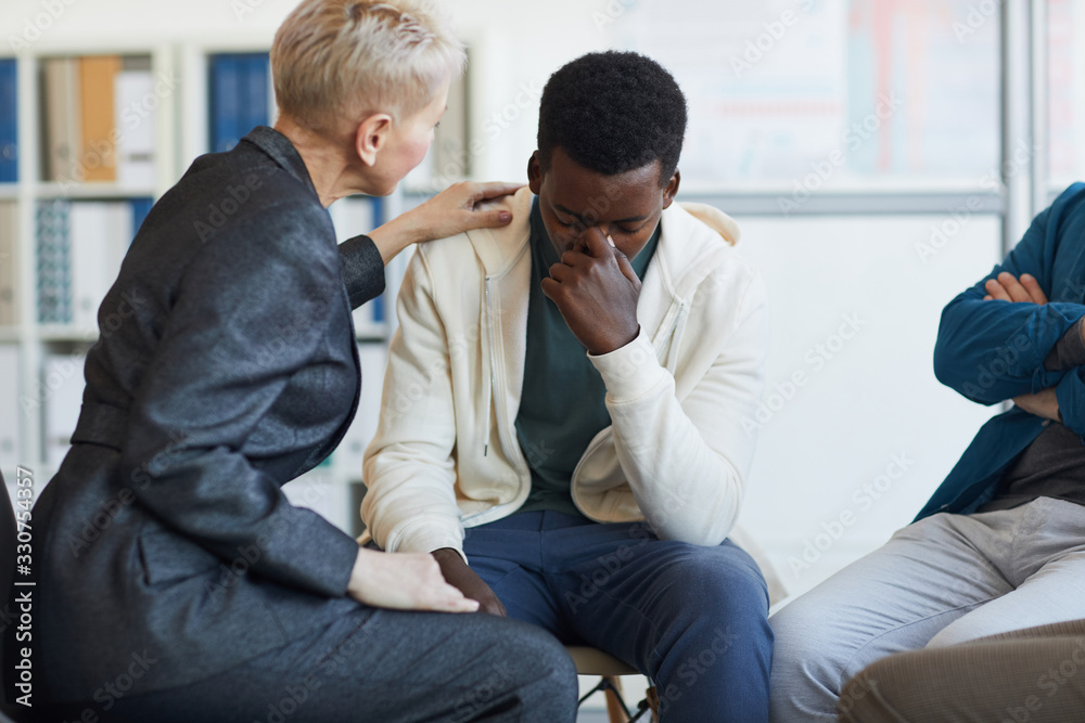 Portrait of young African-American man crying while sitting on chair in support group circle, copy space
