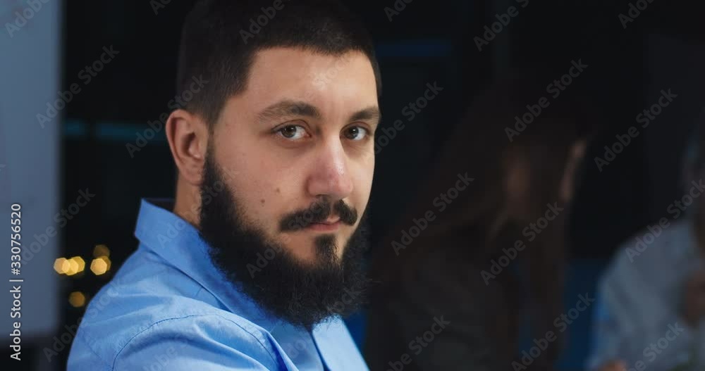 Close up of Caucasian good looking man with beard working at night in office and turning face to camera. Portrait of young businessman looking and smiling late in evening. Close up of Caucasian good looking man with beard working at night in office and turning face to camera. Portrait of young businessman looking and smiling late in evening.