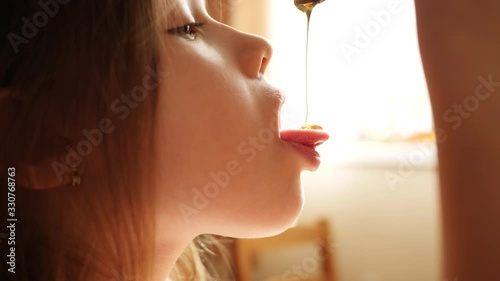 Little child girl eat honey as it drops on her tongue licking it out from the spoon