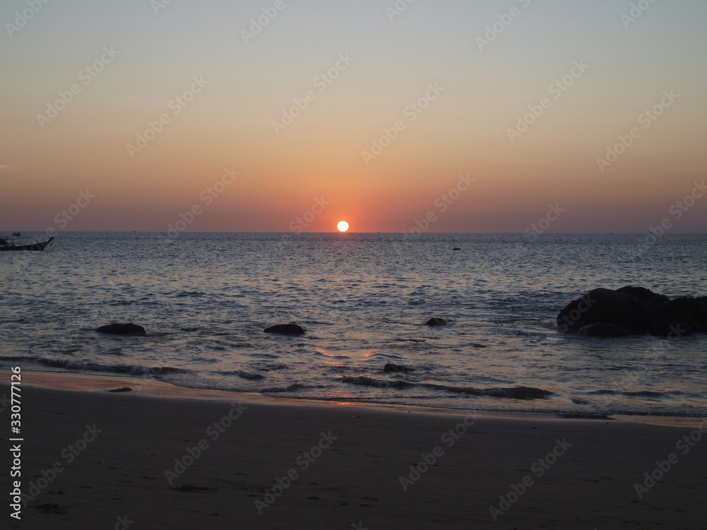 Orange sunset in the sea. In the foreground are stones in the sea, small waves, sand. In the background a ball of the sun moving beyond the horizon, a rock, a longtail boat